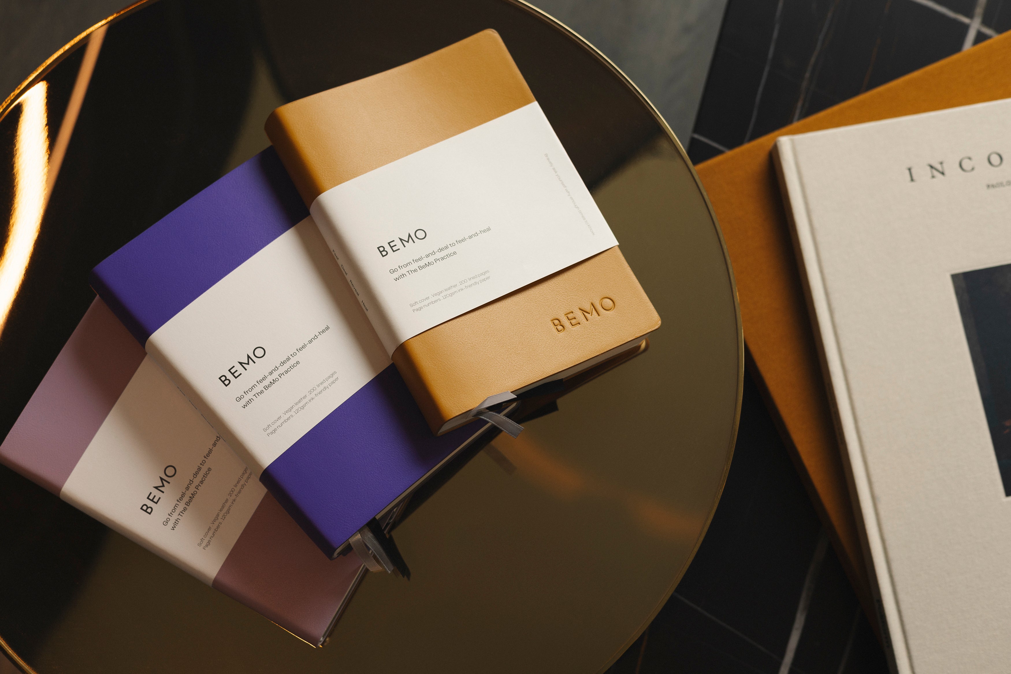 Three BEMO notebooks stacked on a dark surface with a blurred book in the background.